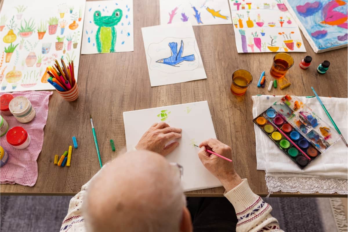 Senior citizen at a wooden table painting on a white sheet of paper with other paintings spread across the table